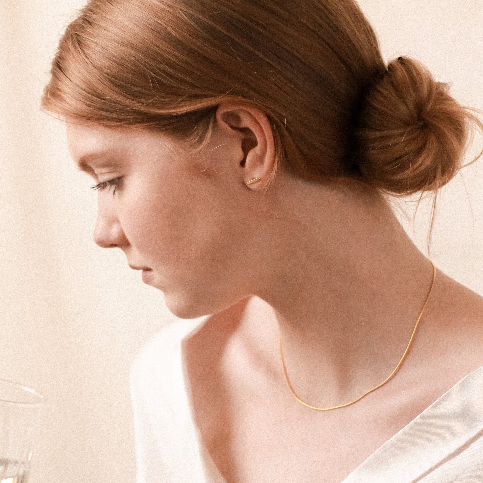 A woman with light brown hair in a low bun wears gold Dainty Bar Studs and a white top, looking down at a glass.