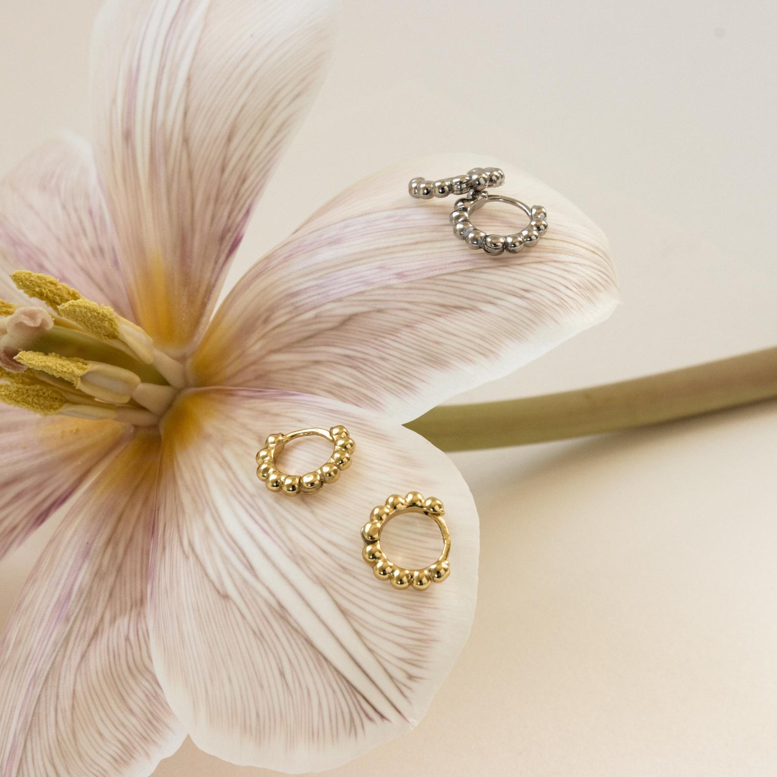 Dainty Sphere Beaded Huggies are shown in gold and silver on a pale flower under soft lighting, highlighting the minimalistic elegance of these hoop earrings.