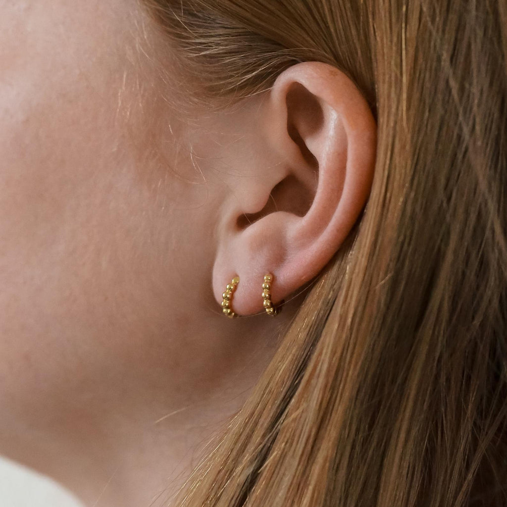 Close-up of a woman’s ear with straight light brown hair, wearing two Dainty Sphere Beaded Huggies—minimalistic small gold hoop earrings.