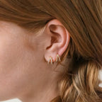 Close-up of a woman's ear wearing the Esther Duo Pave Huggies—V-shaped double hoop gold earrings—with light brown hair pulled back.