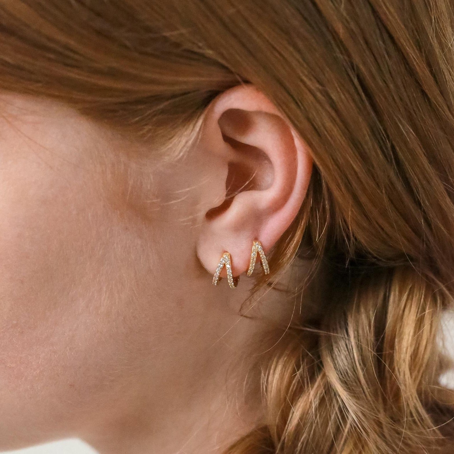 Close-up of a woman's ear wearing the Esther Duo Pave Huggies—V-shaped double hoop gold earrings—with light brown hair pulled back.