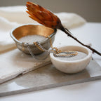 Both gold and silver versions of the necklace draped between two decorative bowls filled with fine white sand.