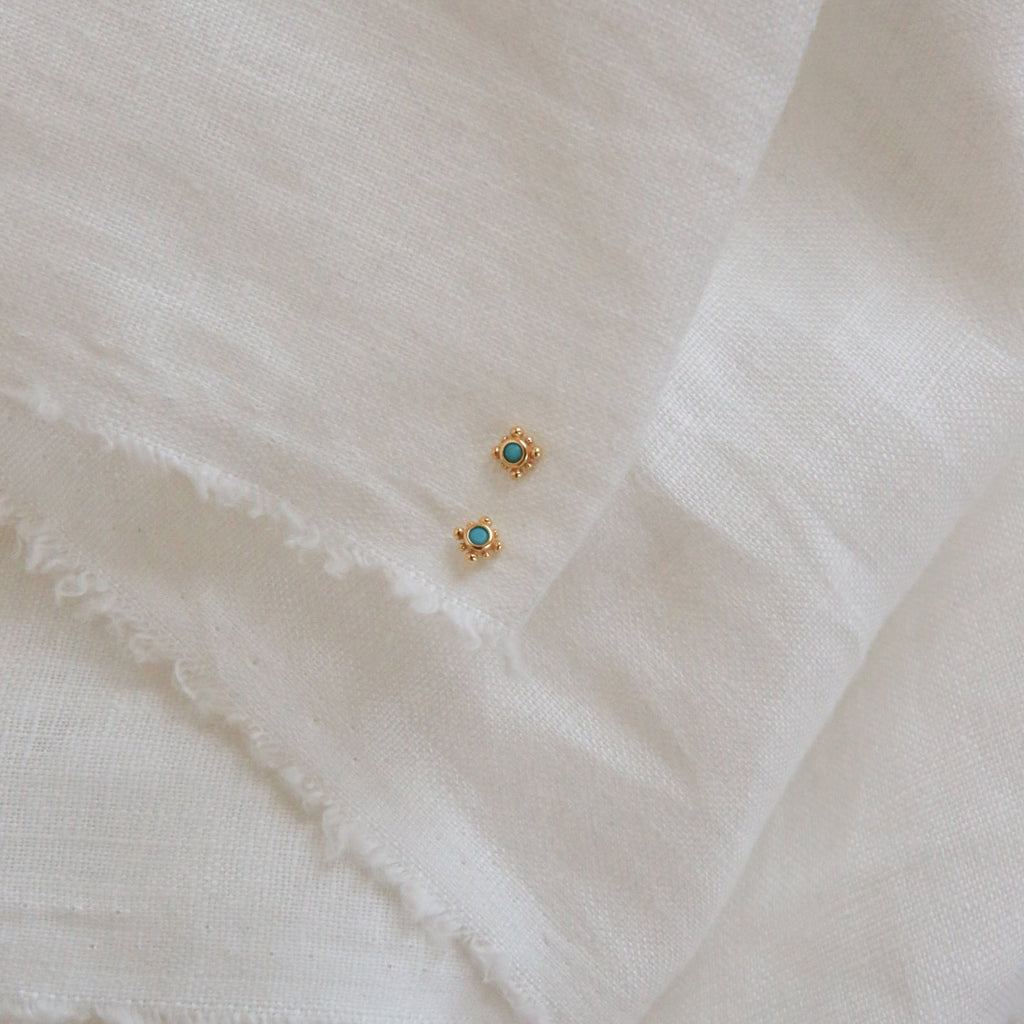 Two Mara Studs, featuring gold and a hint of turquoise, are displayed elegantly on a white cloth.