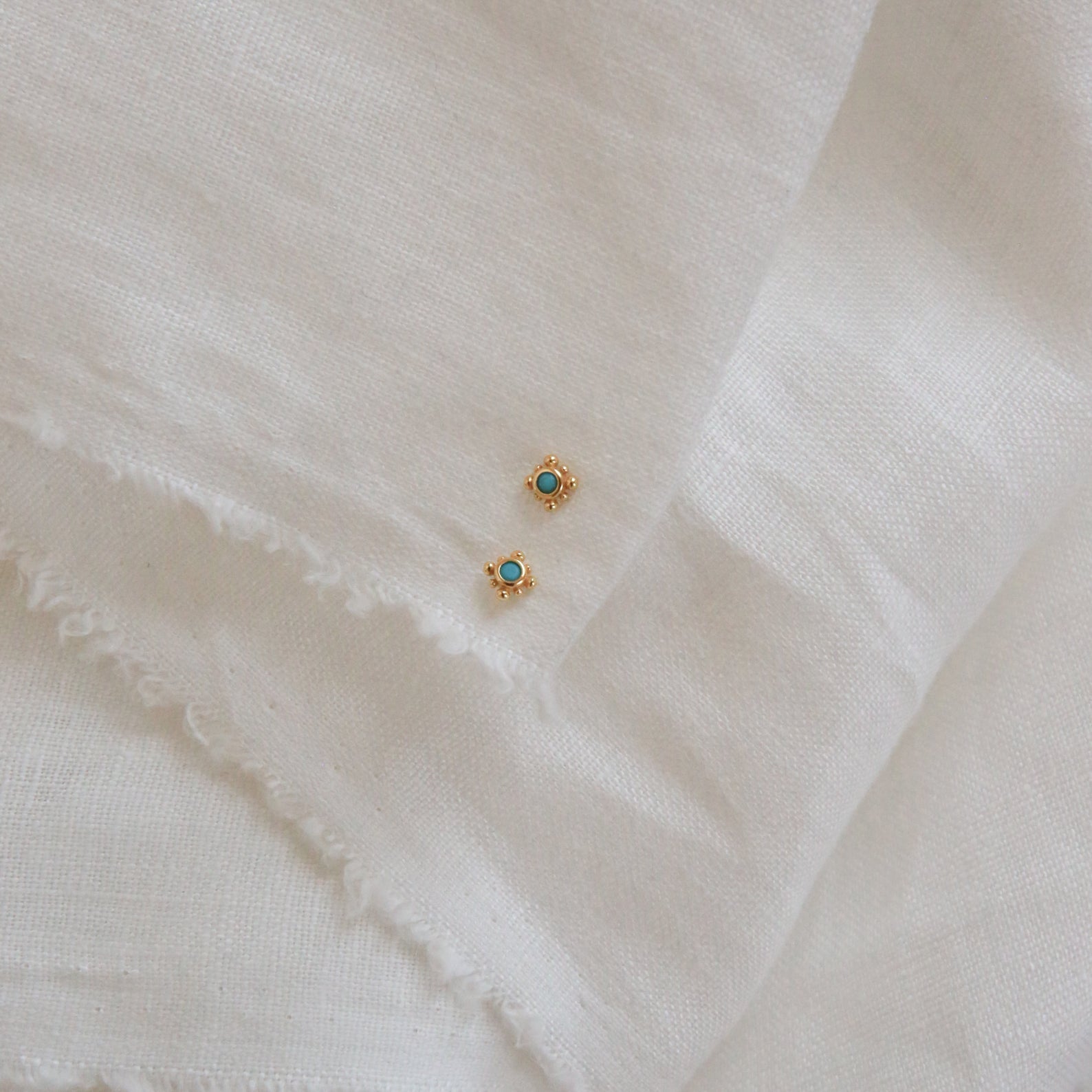 Two Mara Studs, featuring gold and a hint of turquoise, are displayed elegantly on a white cloth.