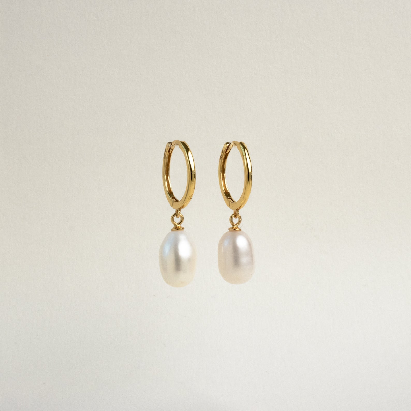 The Maggie Pearl Drop Hoops are gold modern earrings with dangling oval white pearls, displayed against a plain light background—ideal for minimal jewelry lovers.