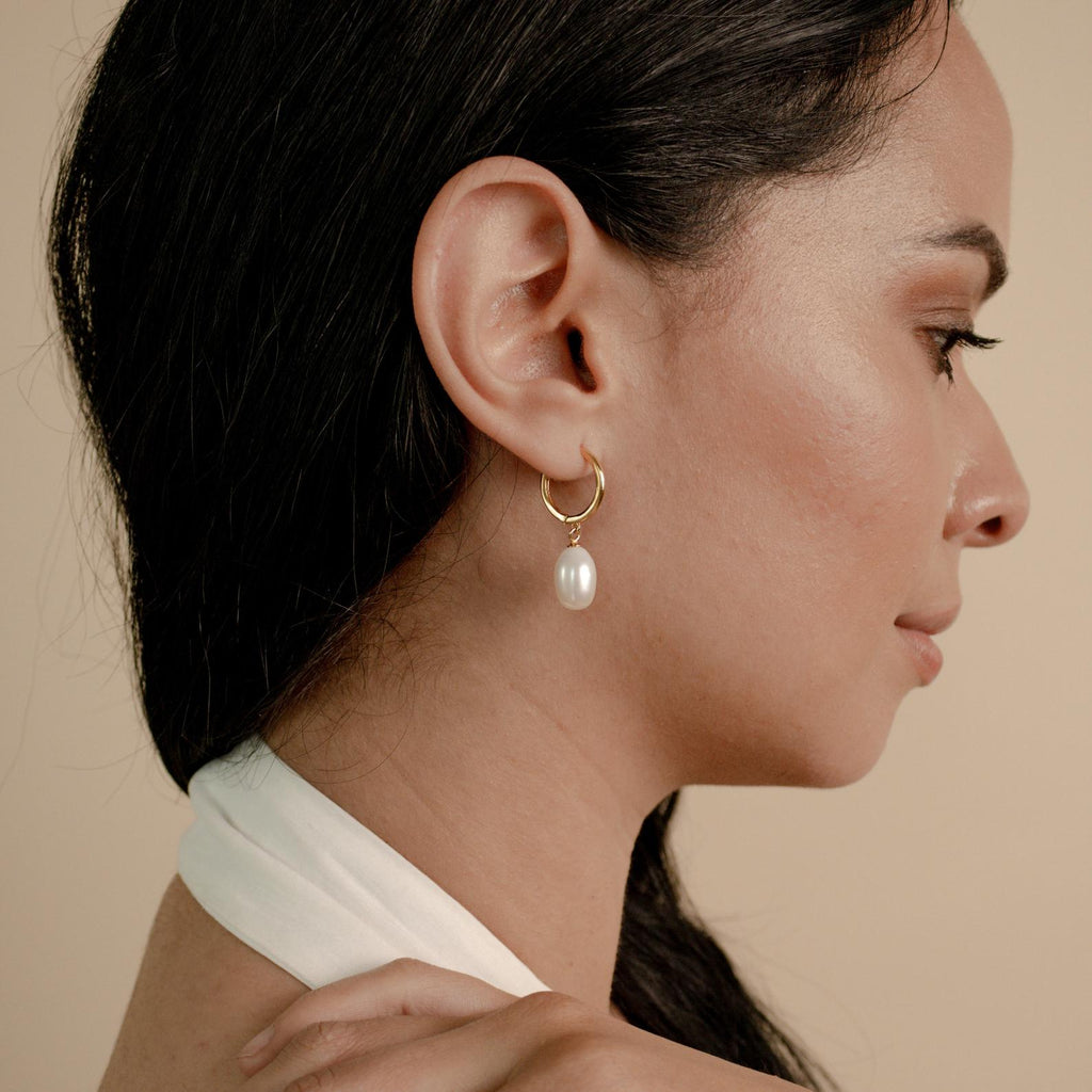 Woman in profile wearing the Maggie Pearl Drop Hoops—modern hoop earrings with a pearl drop—against a beige background.