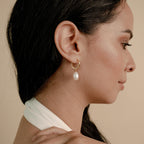 Woman in profile wearing the Maggie Pearl Drop Hoops—modern hoop earrings with a pearl drop—against a beige background.