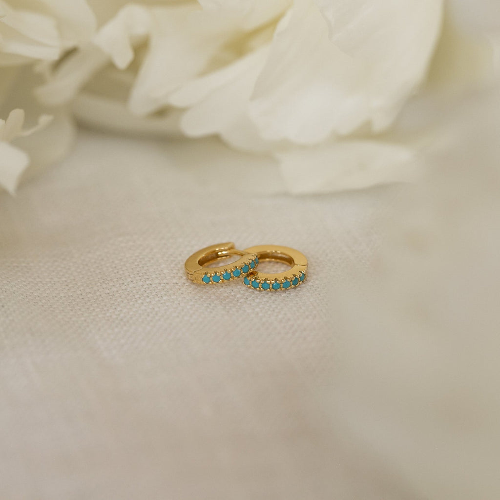 The Kendra Huggies, small gold turquoise earrings with vibrant stones, set on soft white fabric and accented by white flowers, bring eclectic charm to any look.