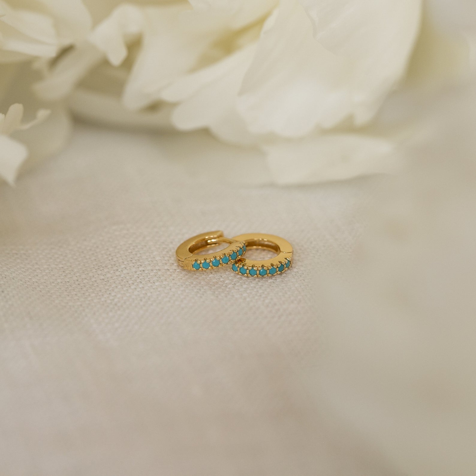 The Kendra Huggies, small gold turquoise earrings with vibrant stones, set on soft white fabric and accented by white flowers, bring eclectic charm to any look.