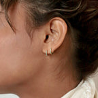 Close-up of a woman's ear wearing two Kendra Huggies, small gold earrings each embellished with green stones on the earlobe for an eclectic touch.
