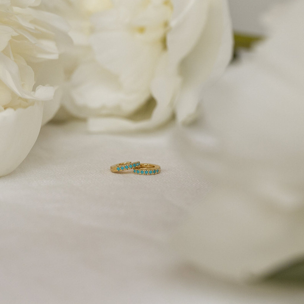 The Kendra Huggies feature gold rings with blue stones, exuding eclectic charm—ideal for stacking or pairing with your favorite turquoise jewelry on a white surface.