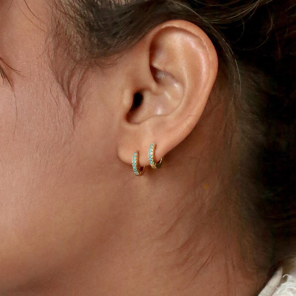 A close-up of a woman's ear wearing Kendra Huggies—two small gold hoops with turquoise stones—that add eclectic charm to her look.