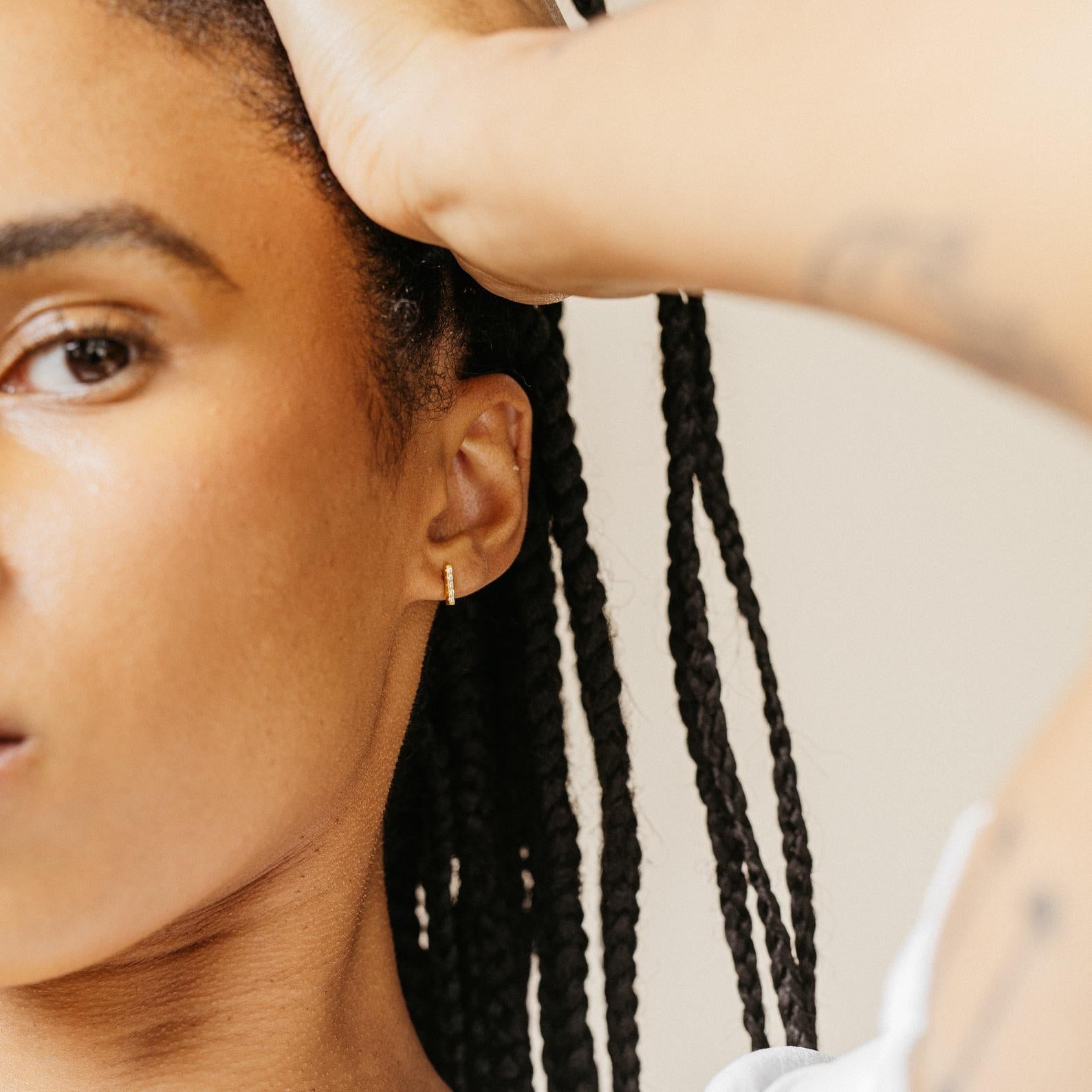A woman with braided hair wears minimal jewelry—Pave Bar Studs—in her ears, her hand resting on her head against a neutral background.