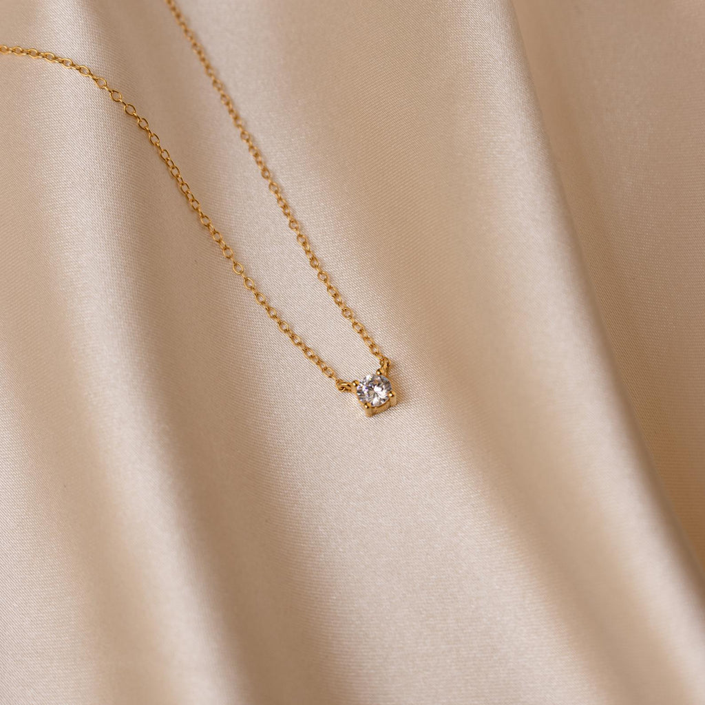 The Dainty Diamond Necklace, featuring a single small diamond charm, is displayed on smooth light beige satin fabric for a soft and elegant presentation.