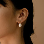 Close-up of a woman wearing Baroque Pearl Earrings with gold studs and large baroque pearl drops, set against a beige background.