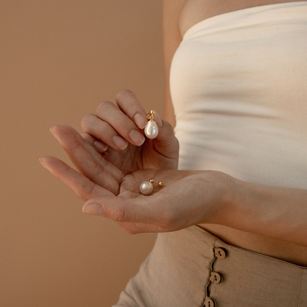 A person in a white tube top holds the Baroque Pearl Earrings with gold hooks, highlighting these statement earrings against a beige background.