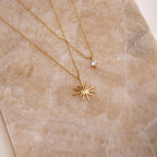 The Laura Starburst Duo Necklace in 18K Gold features two gold necklaces resting on a beige textured surface: one with a round gemstone pendant and the other showcasing a central stone in a starburst design.