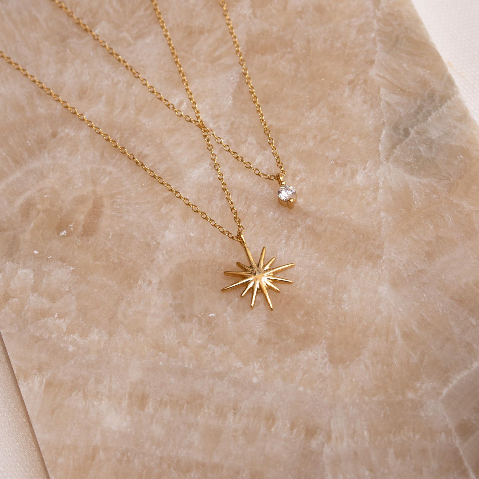The Laura Starburst Duo Necklace in 18K Gold features two gold necklaces resting on a beige textured surface: one with a round gemstone pendant and the other showcasing a central stone in a starburst design.