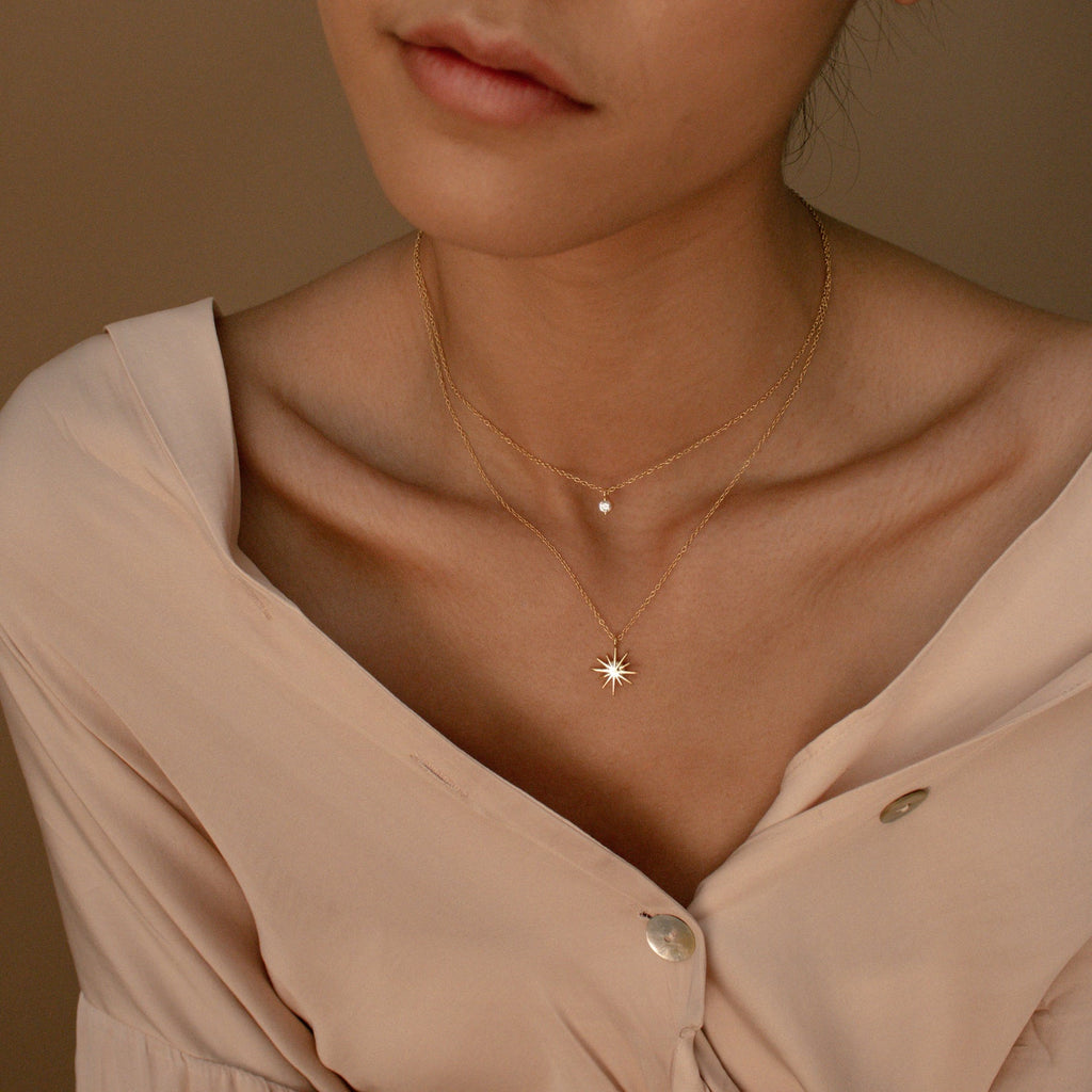 A woman wears a light beige blouse with the Laura Starburst Duo Necklace in 18K Gold, featuring a delicate pendant and a starburst charm on layered chains.