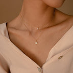 A person wears a beige buttoned blouse and the Laura Starburst Duo Necklace in 18K Gold, featuring two gold chains—one with a small round pendant and the other with a starburst charm.