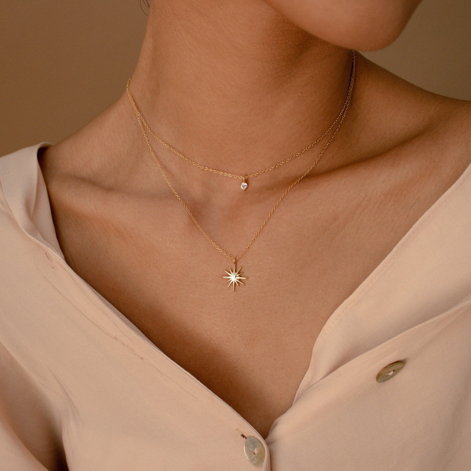 A person wears a beige buttoned blouse and the Laura Starburst Duo Necklace in 18K Gold, featuring two gold chains—one with a small round pendant and the other with a starburst charm.