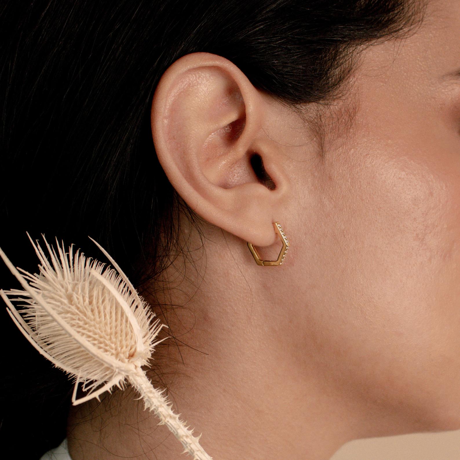 Close-up of a woman's ear wearing Parker Pave Hexagon Huggies, gold geometric earrings, with a dried thistle flower in the foreground.