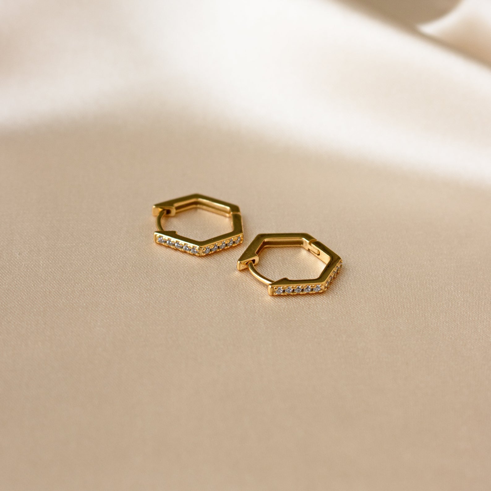 The Parker Pave Hexagon Huggies, crafted in gold and embellished with clear stones, rest on soft beige fabric, highlighting these elegant and stylish pave earrings.