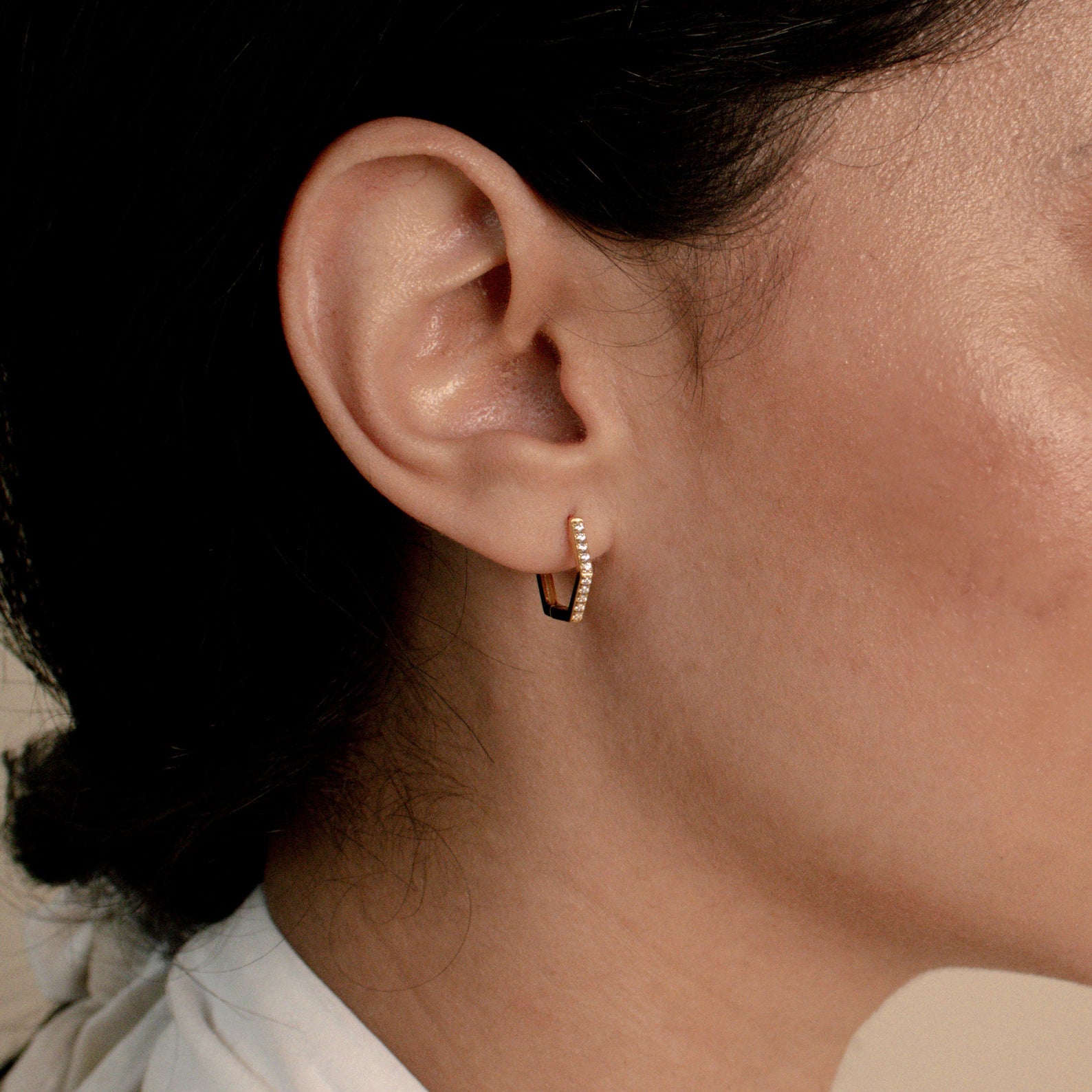 Close-up of a woman's ear wearing Parker Pave Hexagon Huggies, the small gold earrings sparkling with embedded gemstones.