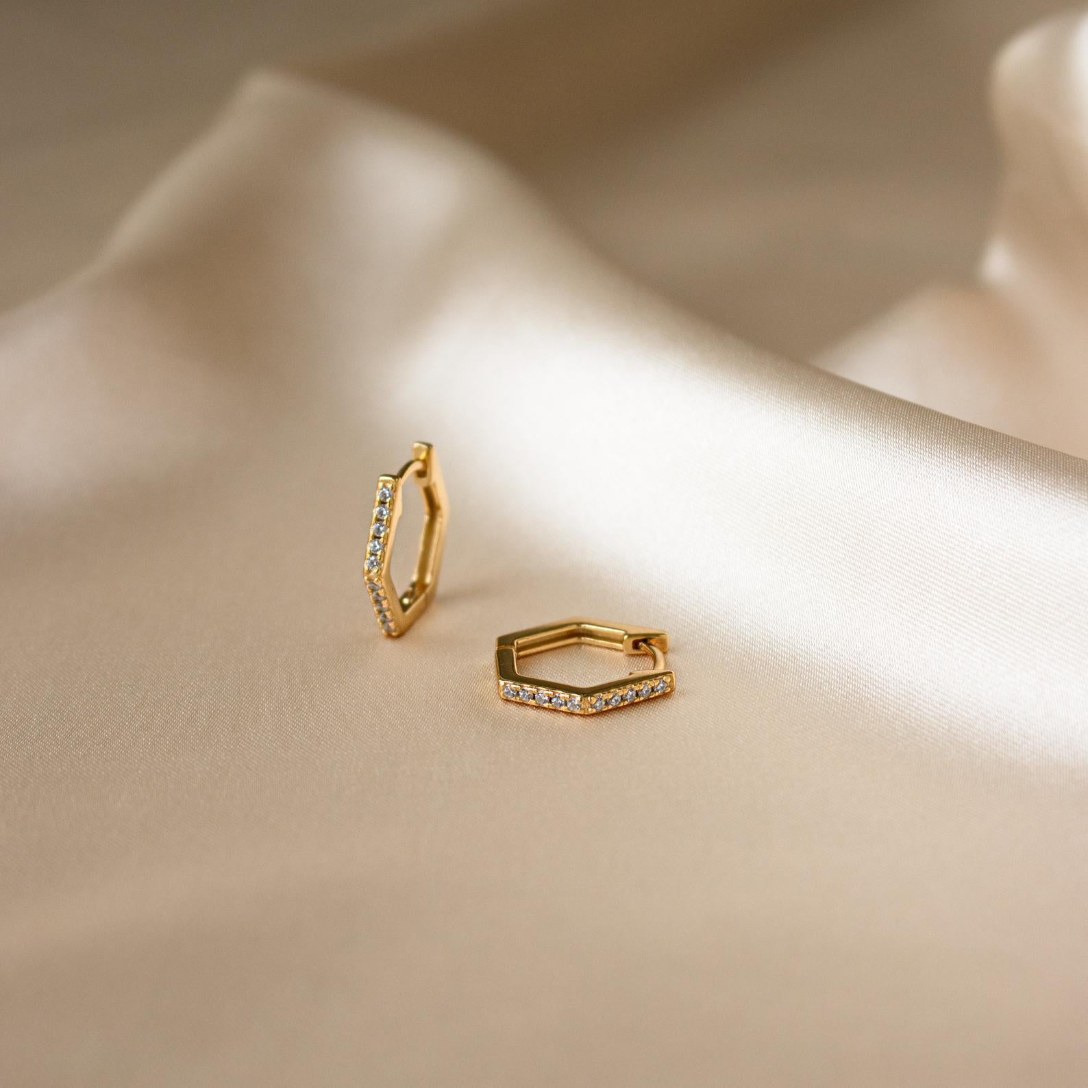 The Parker Pave Hexagon Huggies, chic gold earrings with small crystals, are shown as a stylish ear adornment on smooth champagne-colored satin fabric.