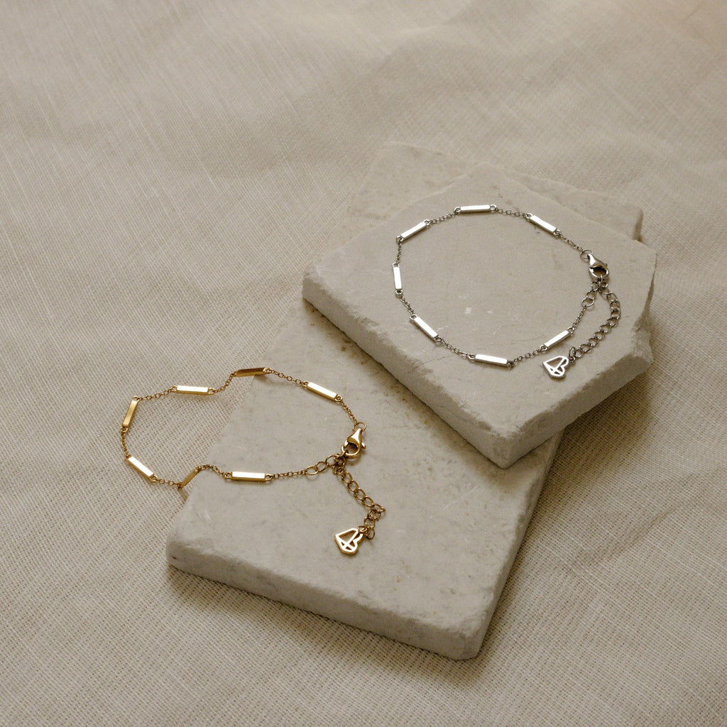 The Abby Bar Link Bracelet is shown in gold and 925 sterling silver on light stone slabs over textured beige fabric, each with a delicate chain and a small heart charm.