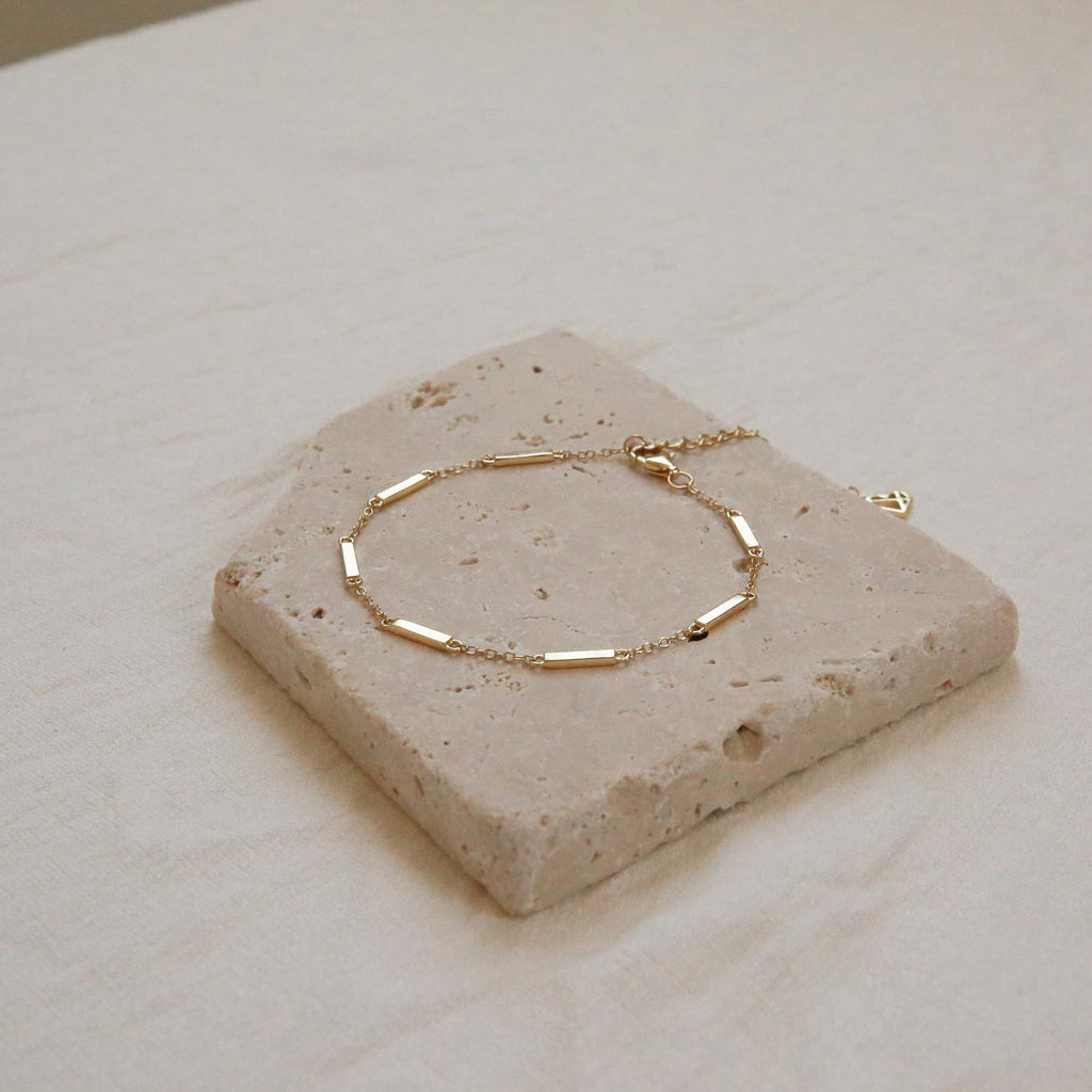 The Abby Bar Link Bracelet, featuring delicate gold rectangular links, is displayed on a square stone tile against a neutral fabric background.