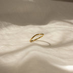 The Dainty Wave Band, a simple and contemporary ring, rests on softly textured cream-colored fabric in gentle sunlight and shadows.