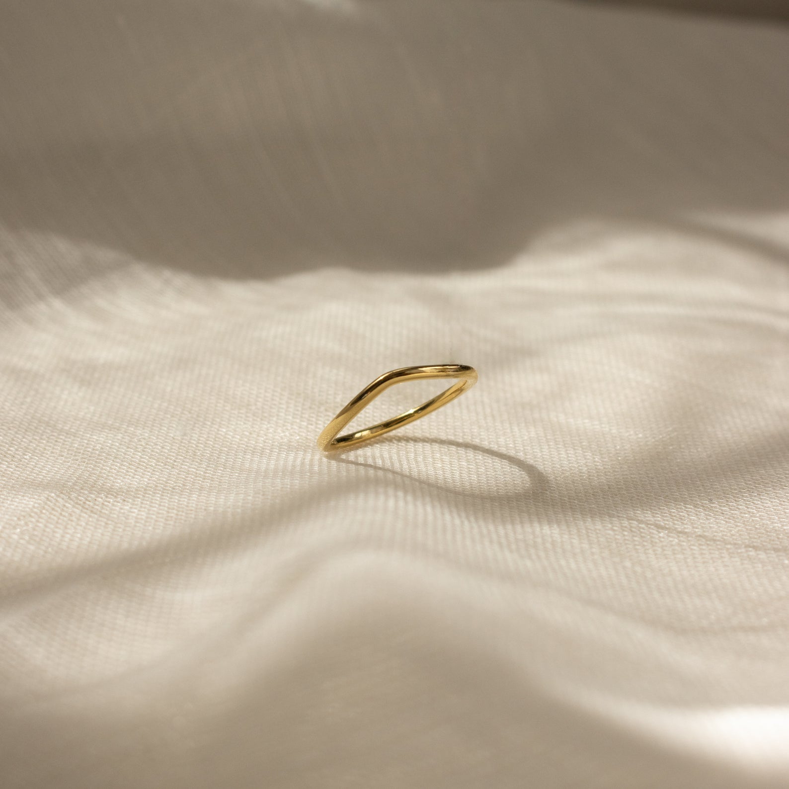 The Dainty Wave Band, a simple and contemporary ring, rests on softly textured cream-colored fabric in gentle sunlight and shadows.
