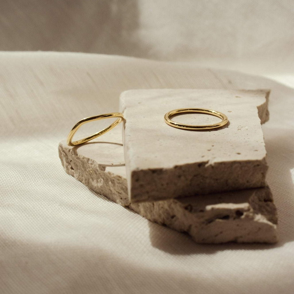 The Dainty Wave Band, a simple gold ring, is displayed on rough, light stone against a soft neutral background—perfect for fans of modern jewelry.