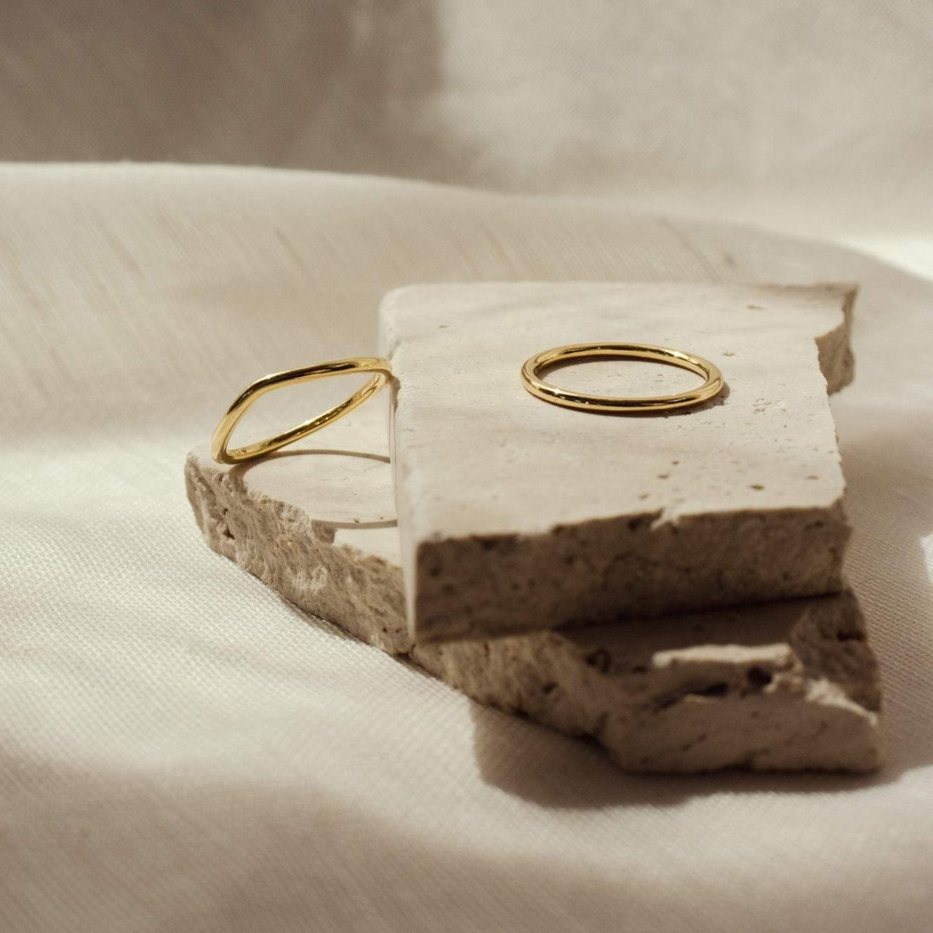 The Dainty Wave Band, a simple gold ring, is displayed on rough, light stone against a soft neutral background—perfect for fans of modern jewelry.