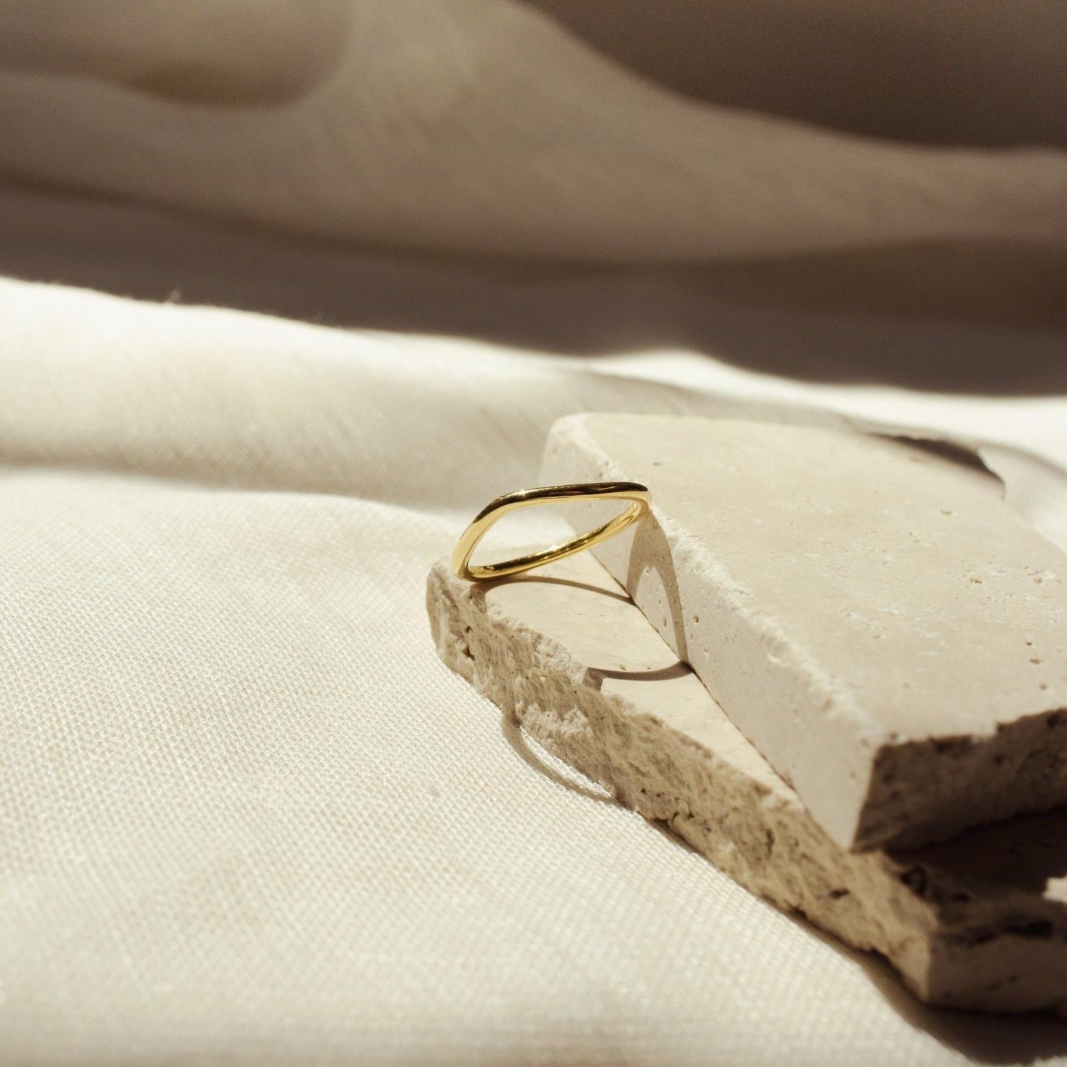 The Dainty Wave Band, a thin gold ring, is displayed on stacked stone slabs over a cream fabric background in gentle sunlight, capturing the essence of modern jewelry elegance.