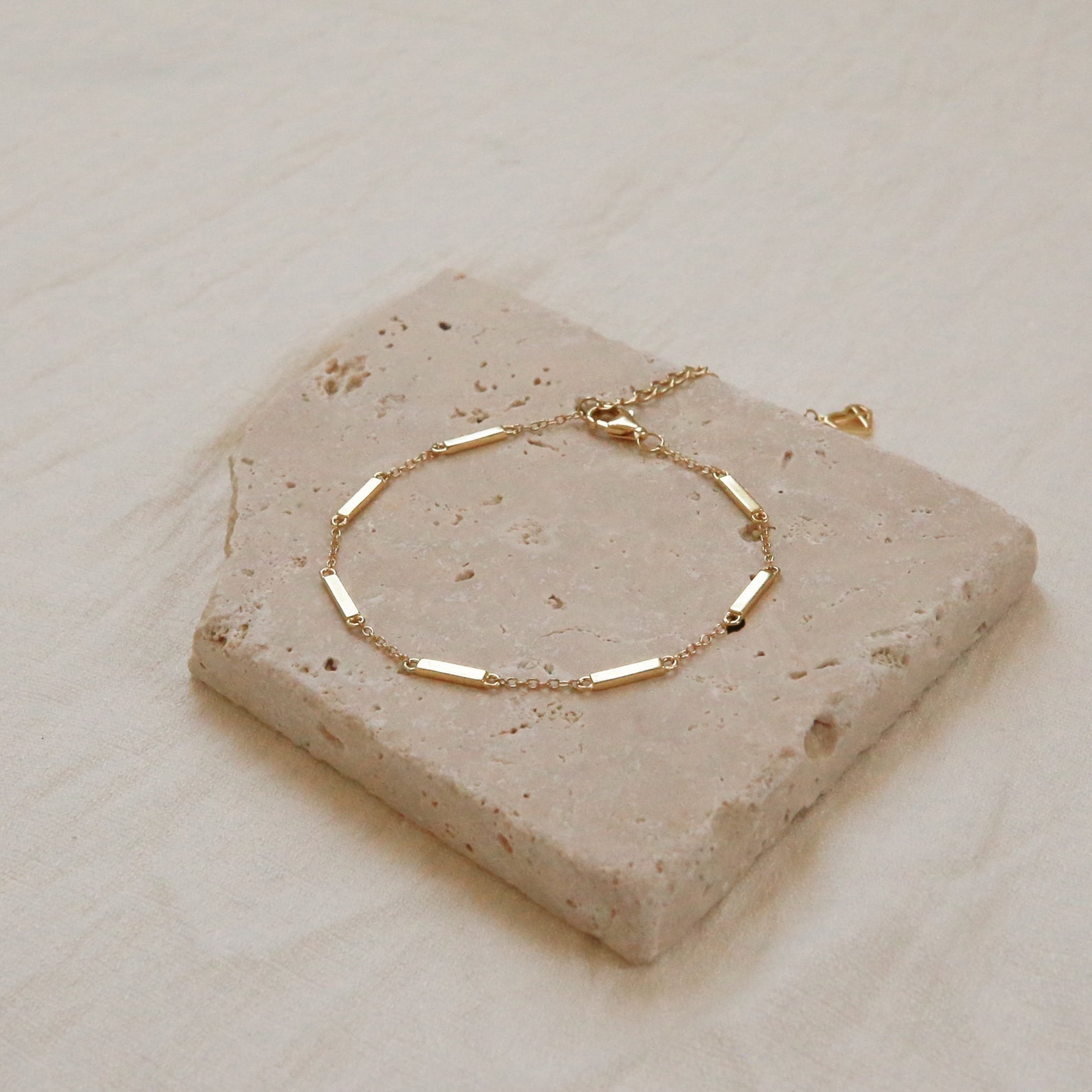 The Abby Bar Link Bracelet is a dainty adjustable gold bracelet with rectangular links, shown on a textured stone tile and beige fabric background.