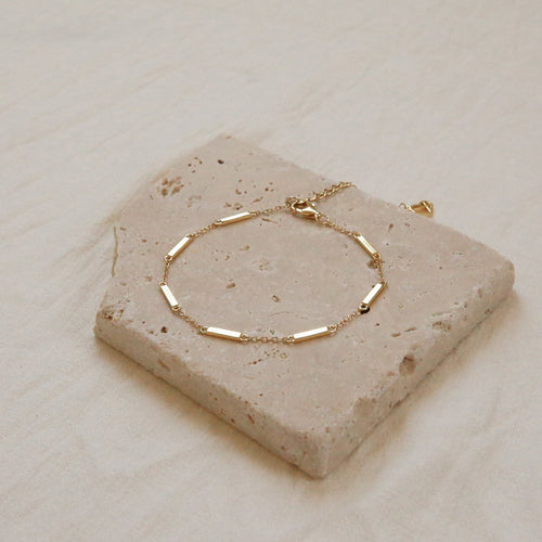 The Abby Bar Link Bracelet is a dainty adjustable gold bracelet with rectangular links, shown on a textured stone tile and beige fabric background.