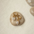 Hazel Pearl Hoops with white pearls rest on a smooth beige stone atop a light fabric surface—an elegant modern jewelry accent.