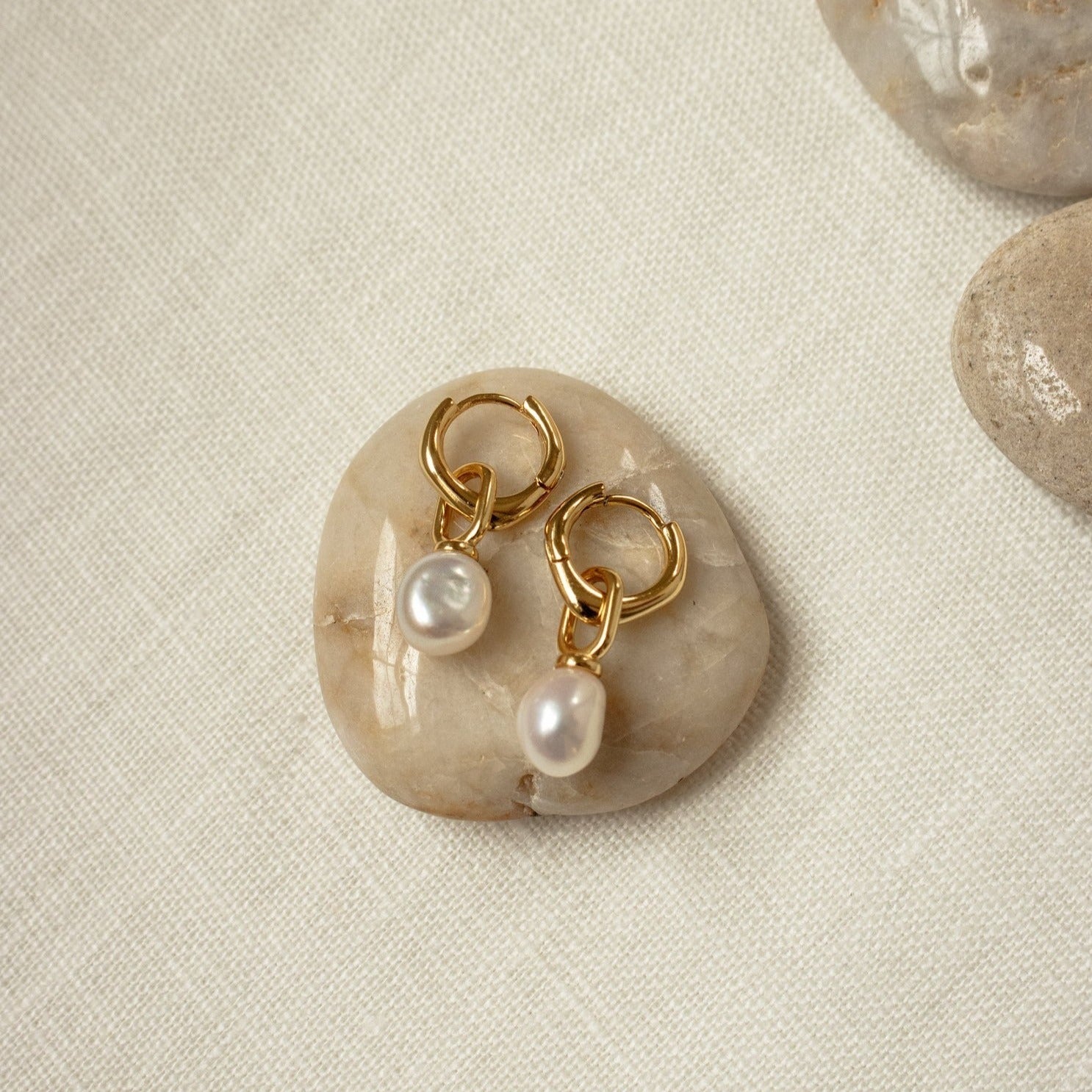 Hazel Pearl Hoops with white pearls rest on a smooth beige stone atop a light fabric surface—an elegant modern jewelry accent.