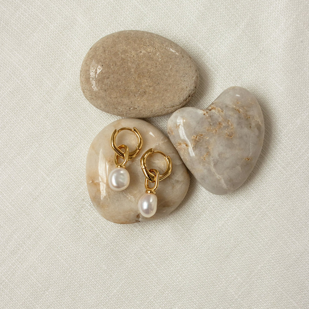 The Hazel Pearl Hoops—gold hoop earrings with pearl drops—are displayed on heart-shaped and smooth beige stones atop white fabric, offering a modern jewelry accent that instantly elevates your style.