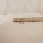 Emerald Diamond Studs featuring rectangular prong-set clear stones, displayed on a light beige stone surface with a minimalist background.