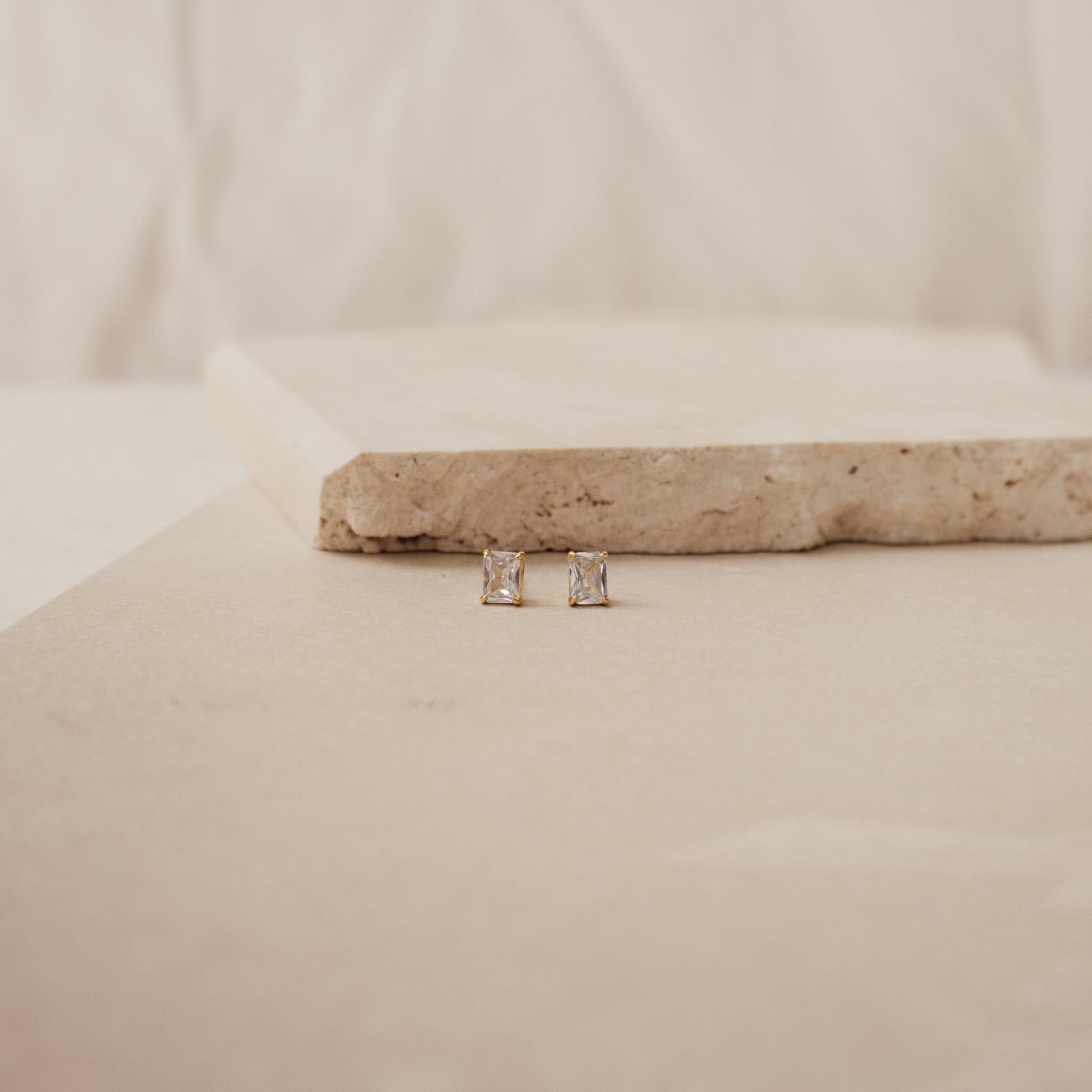 Emerald Diamond Studs featuring rectangular prong-set clear stones, displayed on a light beige stone surface with a minimalist background.