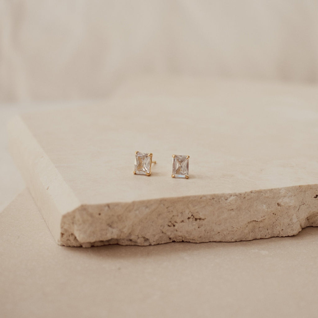 Emerald Diamond Studs with square-cut clear stones in gold prong settings rest on a beige stone surface, set against a soft, neutral background.