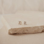 Emerald Diamond Studs with square-cut clear stones in gold prong settings rest on a beige stone surface, set against a soft, neutral background.