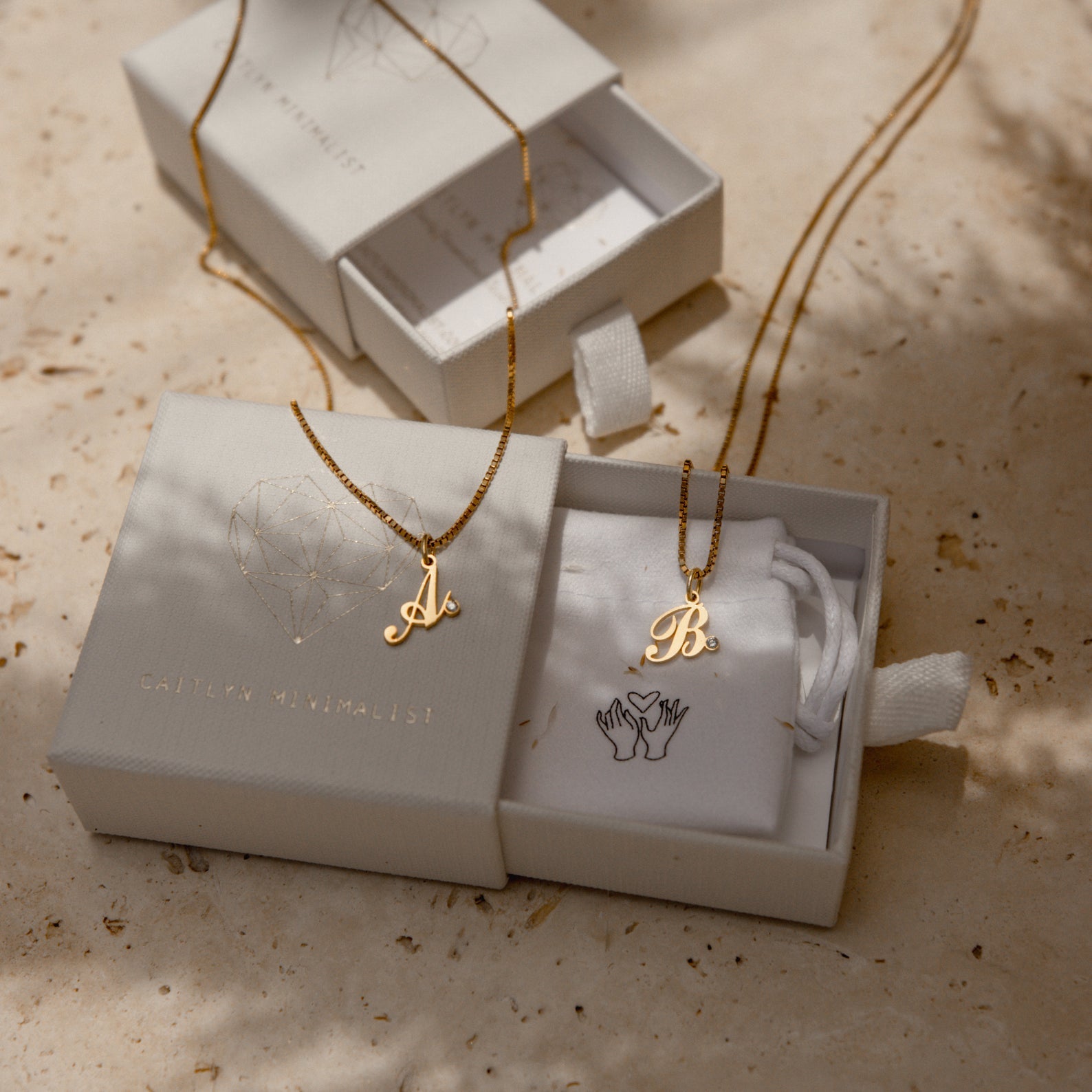 Two Initial Birthstone Necklaces with gold A and B charms in white jewelry boxes on a beige surface—perfect personalized gifts or monogrammed charms for someone special.