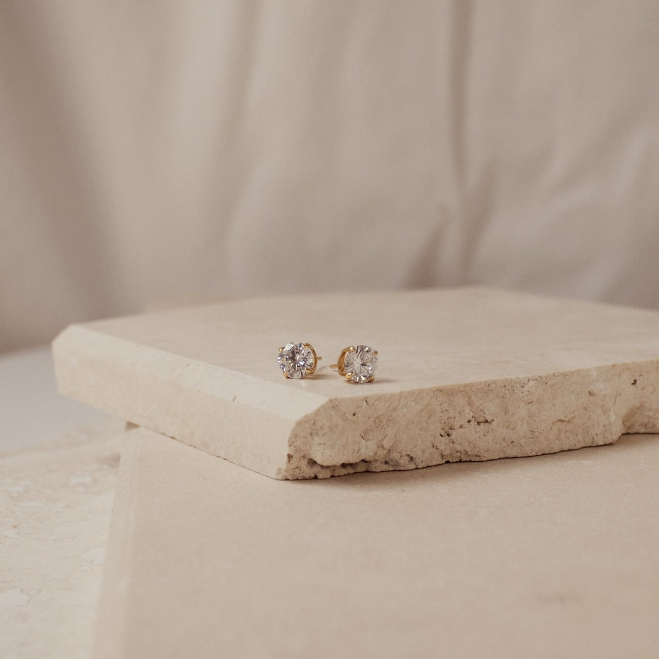 Round Diamond Studs, classic gold earrings featuring round clear gemstones, are displayed on beige stone slabs against a soft background.