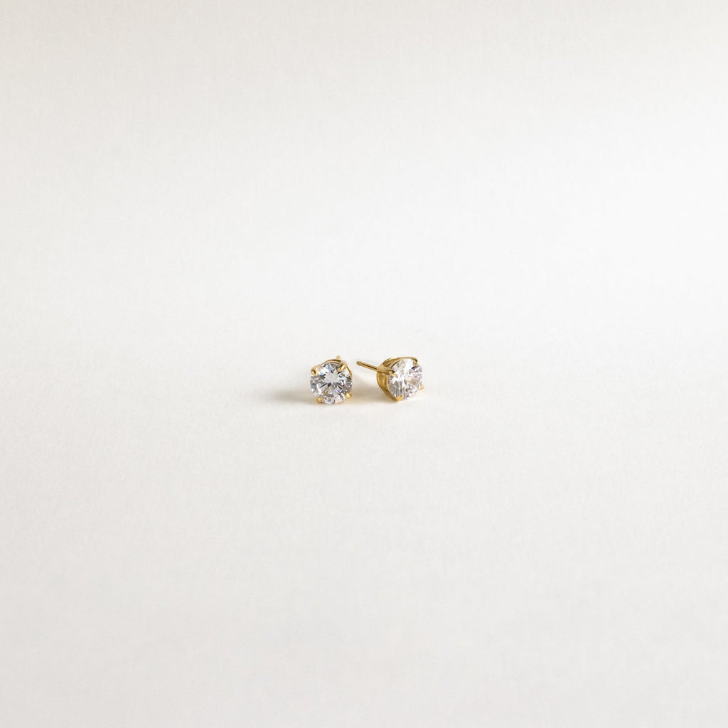 The Round Diamond Studs are classic gold stud earrings featuring round clear gemstones, evoking timeless elegance, displayed on a plain white background.