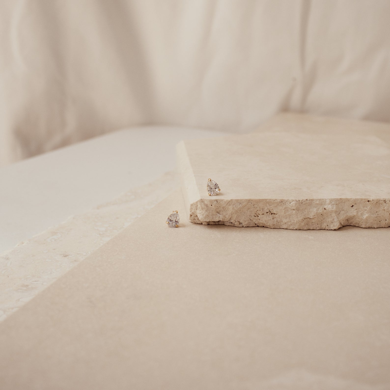 The Pear Diamond Studs, featuring two small, pear-shaped diamonds, rest on a textured beige stone surface with a neutral backdrop—an ideal choice for lovers of minimalistic earrings.