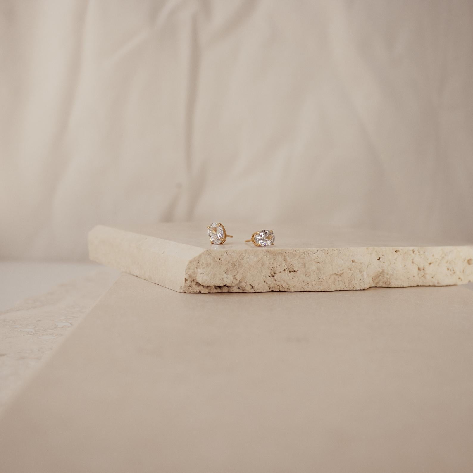 Oval Diamond Studs featuring clear stones are showcased on textured beige stone slabs against a soft background, offering a timeless classic jewelry touch.
